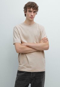 T-shirt beige in cotone con maniche corte e scollo rotondo, indossata da un modello maschile, abbinata a jeans grigio scuro su uno sfondo neutro.