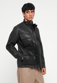 Black leather jacket with a high collar, two front flap pockets, and a zip closure. The texture is smooth with a slight sheen.