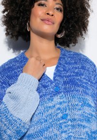 Niebieski i jasnoniebieski dziergany kardigan z dekoltem w serek, długimi rękawami oraz dwoma niebieskimi guzikami. Teksturowana przędza nadaje mu miękki, przytulny wygląd.