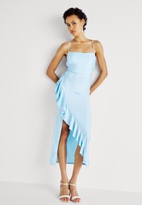 Robe bleu clair avec des bretelles spaghetti, buste ajusté et ourlet asymétrique avec des volants. Texture douce avec une fente sur le côté, assortie à des talons blancs.