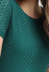 Tyrkysový pletený top s texturovaným vzorem a volutovými detaily. Krátké rukávy a kulatý výstřih.