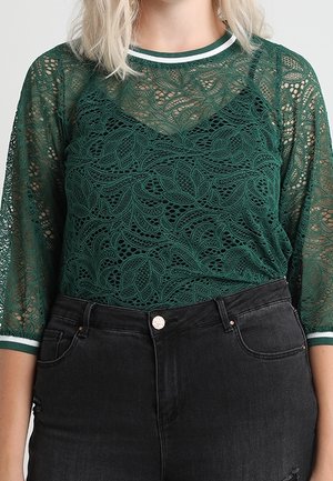 Kobieta ubrana w ciemnozielony, kwiatowy top z koronki z rękawami do połowy przedramienia i paskowanym dekoltem, w połączeniu z czarnymi, wysokim stanem jeansami.
