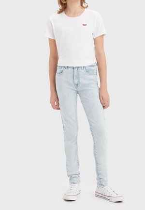 Person iført en hvid kortærmet cropped t-shirt med et lille rødt logo, lyseblå skinny jeans og hvide højt opskårne sneakers.