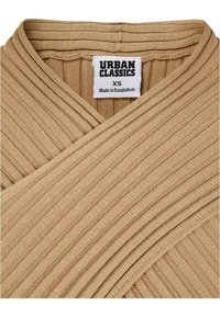 Tissu côtelé beige avec un design en V. L'étiquette indique "URBAN CLASSICS, XS, Fabriqué au Bangladesh." La texture semble légèrement rugueuse.
