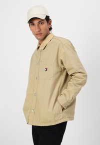 Beige knapjacke med en cordfløjlskrage, brystlomme og logo patch. Modellen bærer også en lysfarvet kasket.