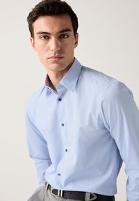 Ljusblå skjorta med knappslå och texturerat mönster, kort krage och kontrasterande innerkrage. Bärs med mörkt bälte och grå byxor.