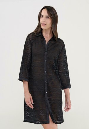 Mujer vestida con un vestido negro de manga larga abotonado, con un patrón geométrico de encaje, de pie frente a un fondo liso gris claro.