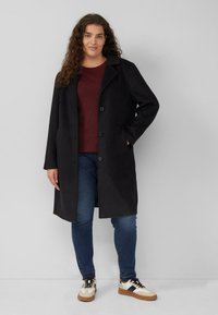 Zwarte wollen blend jas met een notch-kraag, drie knopen en zijzakken. Gecombineerd met een bordeauxrode trui, blauwe jeans en witte sneakers.