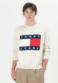Maglione crema con un logo rettangolare con "TOMMY" e "JEANS" in blu navy, rosso e bianco. Maniche e orlo a coste, design classico a girocollo.