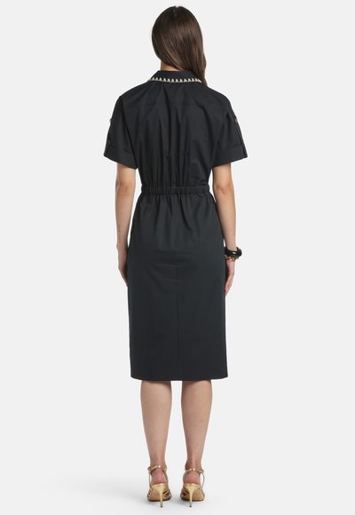 Žena v čiernych krátkych rukávoch, šaty po kolená s elastickým pásom a zlatými vysokými sandálmi na opätku, zobrazená zozadu.