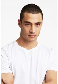 Valkoinen pyöreäkauluksinen t-paita miehen mallilla, jolla on lyhyet tummat hiukset. T-paidassa on sileä tekstuuri ja se on istuva, ja taustana on yksinkertainen tausta.