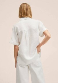 Chemise en lin blanche avec un col à revers, des manches courtes retroussées et un pli au dos, associée à un pantalon assorti de couleur claire.