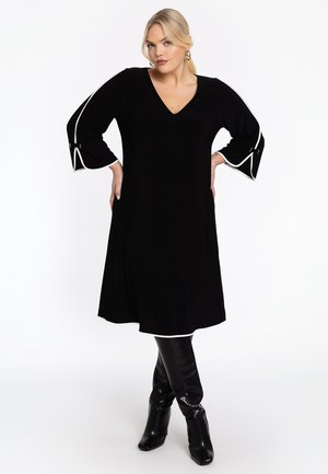 Robe noire avec un col en V, manches cloche à trois quarts avec des accents blancs. Tissu lisse, longueur genoux, associée à des bottes à talons noires.