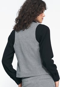 Czarna koszula z długim rękawem w parze z szarym kamizelką w wzór jodełki. Kamizelka ma skrócony krój i detal w postaci wentylacji z tyłu.