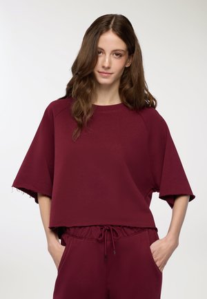 Giovane donna con lunghi capelli castani che indossa una maglietta corta e ampia color maroon e pantaloni abbinati con cordini, mani nelle tasche, sfondo neutro.