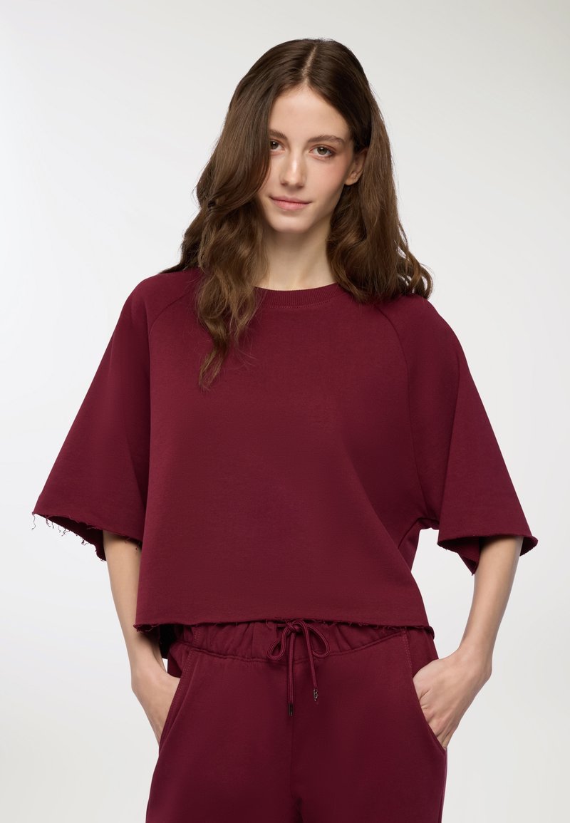 Giovane donna con lunghi capelli castani che indossa una maglietta corta e ampia color maroon e pantaloni abbinati con cordini, mani nelle tasche, sfondo neutro.