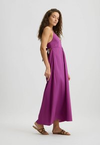 Purple maxi dress with a halter neck, tied at the back, featuring a flowy silhouette and side cutouts. Worn with animal print slip-on sandals.