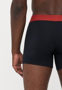 Svarte bomullsboxershorts med rød elastisk midjebånd, som viser bakvinkelen og fremhever sømdetaljene.