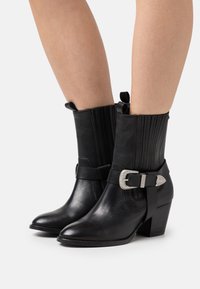 Botas de tobillo de cuero negro con punta afilada, paneles laterales texturizados y un acento de hebilla plateada en una correa. Tacón grueso para mayor estabilidad.