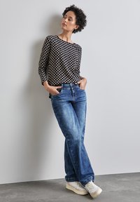 Musta ja beige kuvioitu pitkähihainen paita yhdistettynä vaaleansinisiin farkkuihin ja valkoisiin lenkkareihin, seisoo neutraalin taustan edessä.