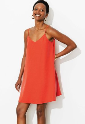 Femme souriante portant une robe orange sans manches et des boucles d'oreilles créoles dorées, debout avec une main sur la hanche devant un fond blanc.