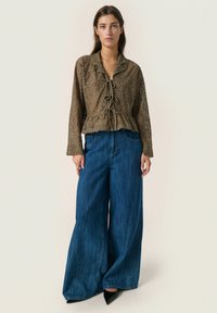 Lace blouse in olive green with a ruffled hem and tied front. Paired with loose, wide-leg blue denim jeans and pointed black shoes.