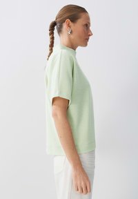 Top corto a maniche corte di un verde chiaro e texturizzato con colletto alto. Presenta una vestibilità ampia e orlo cucito, abbinato a pantaloni bianchi.