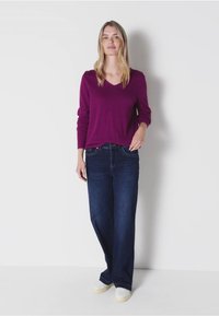 Haut violet à manches longues en V associé à un jean large bleu foncé. Le modèle porte des baskets blanches et se tient devant un fond uni.