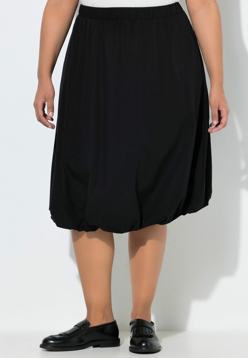 Mujer con una falda negra con cintura elástica, longitud hasta la rodilla y estilo burbuja, combinada con mocasines de cuero negro, sobre un fondo blanco liso.