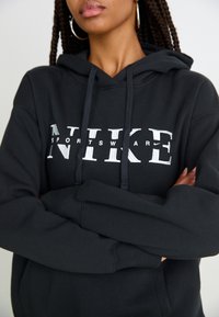 Crna Nike dukserica s prednjim logotipom u bijeloj boji. Ima kapuljaču s podesivim šniranjem i rebraste rukave. Čini se da je materijal mekan i deblji.