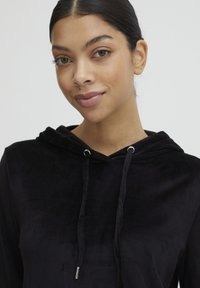 Schwarzer Kapuzenpullover aus weichem, strukturiertem Stoff. Mit silbernen Ösen und Kordeln ausgestattet, hat er eine lockere Passform und lange Ärmel.