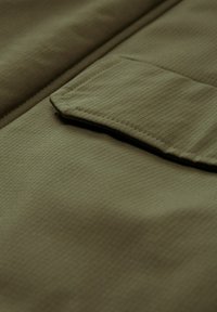 Tessuto verde oliva con una superficie testurizzata, caratterizzato da una tasca quadrata con un'ala inclinata. Il materiale appare liscio e leggermente lucido.