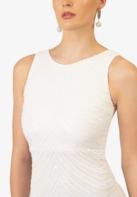 Abito bianco con un motivo a onde tridimensionale, senza maniche e con scollatura a forma di onda. Il modello indossa orecchini a goccia in perle come accessorio.