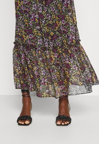 Robe noire à fleurs avec des fleurs violettes, jaunes et roses, dotée d'un ourlet à volants et d'un tissu transparent. Sandales à talons hauts noires tressées.