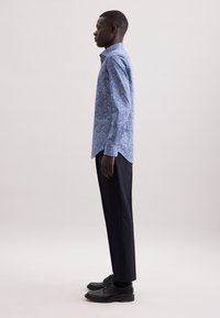 Camicia a maniche lunghe con motivo blu, colletto alla francese e orlo curvo, abbinata a pantaloni scuri e scarpe nere.