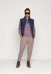 Marineblauw gewatteerd vest over een lichtpaarse top met lange mouwen, gecombineerd met beige taps toelopende broek en bruine wandelschoenen. Zonnebril maakt de look compleet.