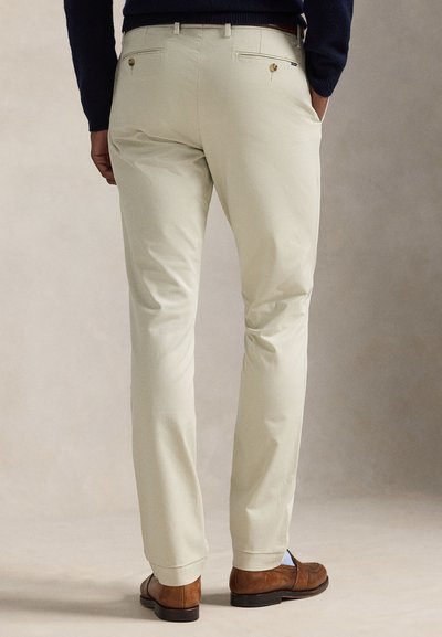 Muž oblečený v béžových úzkych nohaviciach, hnedej koženej mokasíne a tmavom svetri, stojaci s rukami v kapsách na neutrálnom pozadí.