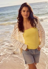 Cardigan léger en tricot crème, haut sans manches jaune et shorts en denim. Texture douce et coupe décontractée, debout sur la plage.