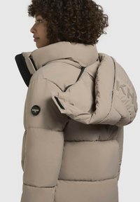 Gaišbrūns pūkains mētelis ar augstu apkakli, lielu kapuci un redzamām šuvēm. Tam ir apaļš logotips un rāvējslēdzēja kabata uz rokas.