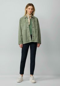 Veste en denim vert avec des boutons et deux poches poitrine, associée à un pantalon sombre et des baskets compensées blanches, agrémentée d'une écharpe légère.