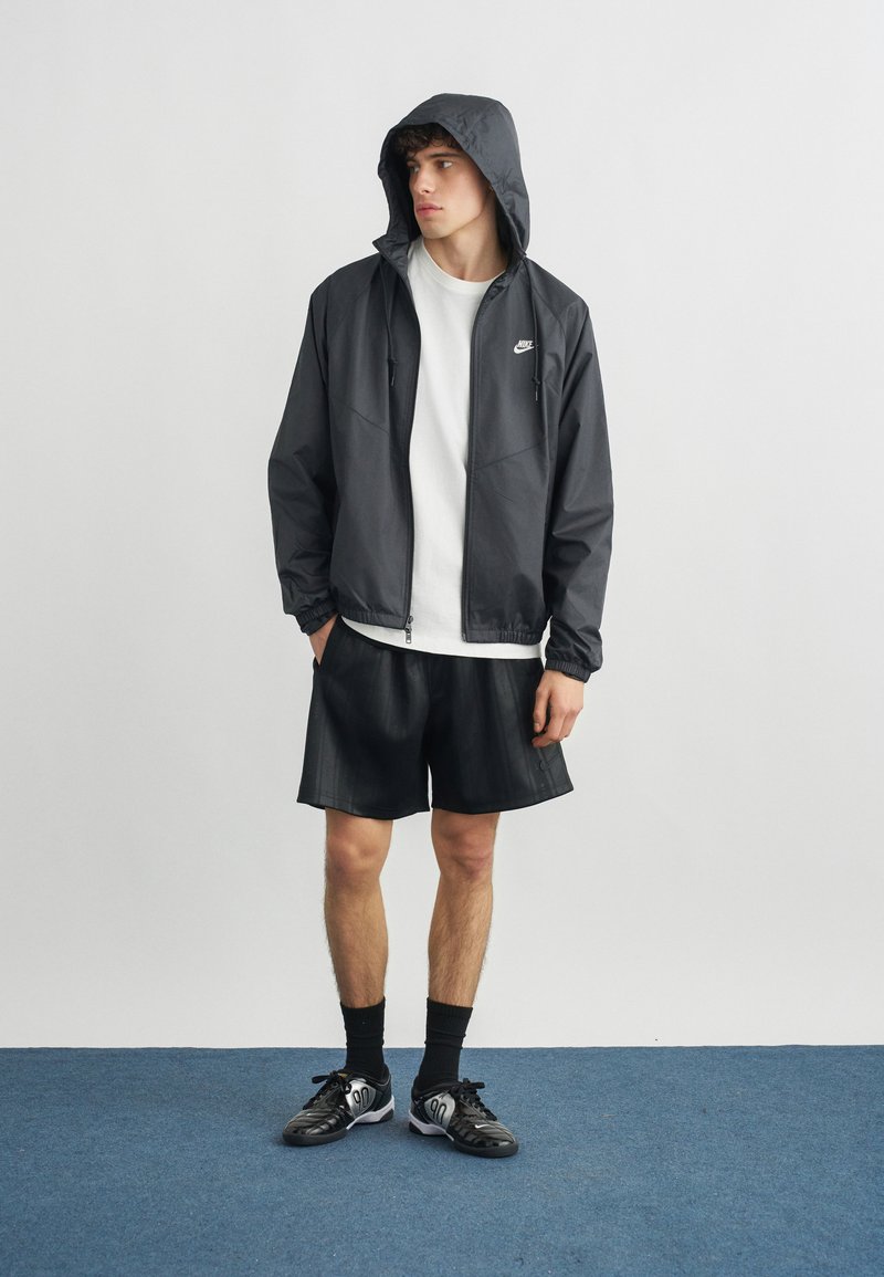 Giovane uomo che indossa una giacca con cappuccio nera Nike, maglietta bianca, pantaloncini neri, calzini neri e scarpe da ginnastica nere in piedi su un tappeto blu.