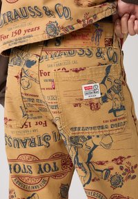 Pantalones de Levi's con temática de Pixar, tejido beige con diseños impresos en rojo y azul, incluyendo personajes y logotipos. Presenta bolsillos laterales y etiqueta.
