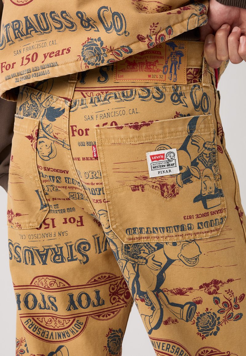 Levi's byxor med Pixar-tema, beige tyg med röda och blå tryckta mönster, inklusive karaktärer och logotyper. Har sidofickor och etikett.