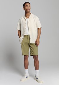 Lichtbeige overhemd met korte mouwen, gecombineerd met olijfgroene shorts. Witte tanktop eronder en witte sneakers met blauwe accenten.
