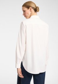 Weißes Langarmhemd mit klassischem Kragen, glatter Textur und abgerundetem Saum, getragen mit dunkelblauen Jeans. Details umfassen geknöpfte Manschetten.