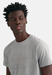 T-shirt en coton gris avec un logo de marque texturé en gris clair. Manches courtes et encolure ronde, près du corps.