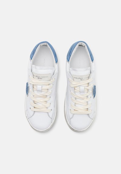 Weiße Sneakers mit blauen Akzenten, ausgestattet mit einem glatten Lederobermaterial, flachen cremefarbenen Schnürsenkeln und einem Logo an der Seite. Abgerundete Zehenform.