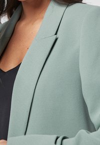 Donna che indossa un blazer verde chiaro sopra una maglietta nera, inquadrata dal petto al collo, con pelle e ciocche di capelli visibili.