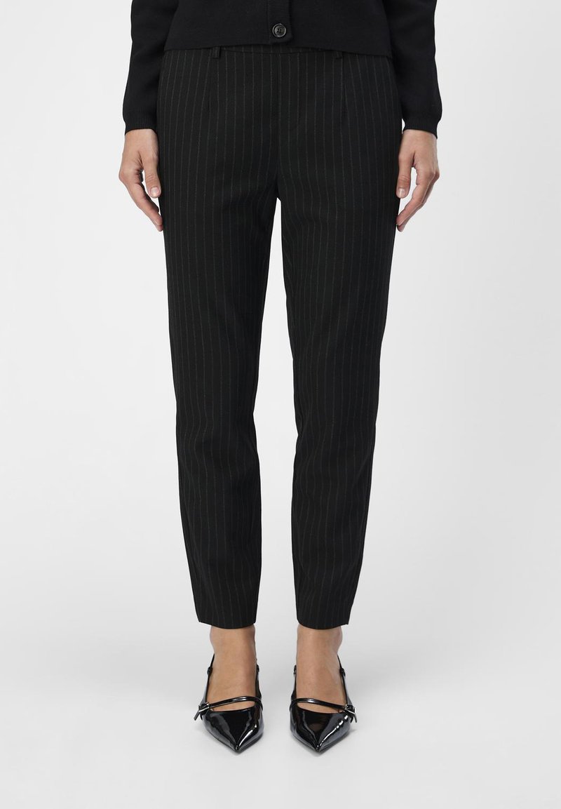 Un pantalon noir à rayures fines présente une coupe ajustée avec une texture lisse. Associé à des escarpins noirs à bout pointu brillants, mettant en valeur des lignes élégantes.