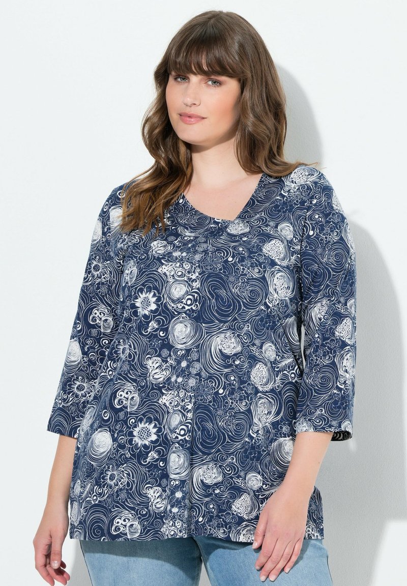 Frau mit braunem, welligem Haar, die ein navyblaues und weißes Oberteil mit floralen Wirbelmustern und drei Viertelärmeln sowie hellblauen Jeans trägt.